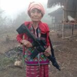 July 2022—In the remote mountains of Chin State, Myanmar, a Chin ethnic woman stands guard, gripping a makeshift r!fle as she watches for approaching danger.